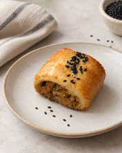 A golden-brown baked pastry or roll topped with black sesame seeds, served on a light beige ceramic plate. The plate is set on a light stone surface with a striped cloth napkin, scattered sesame seeds, and a small bowl of black seeds in the background.