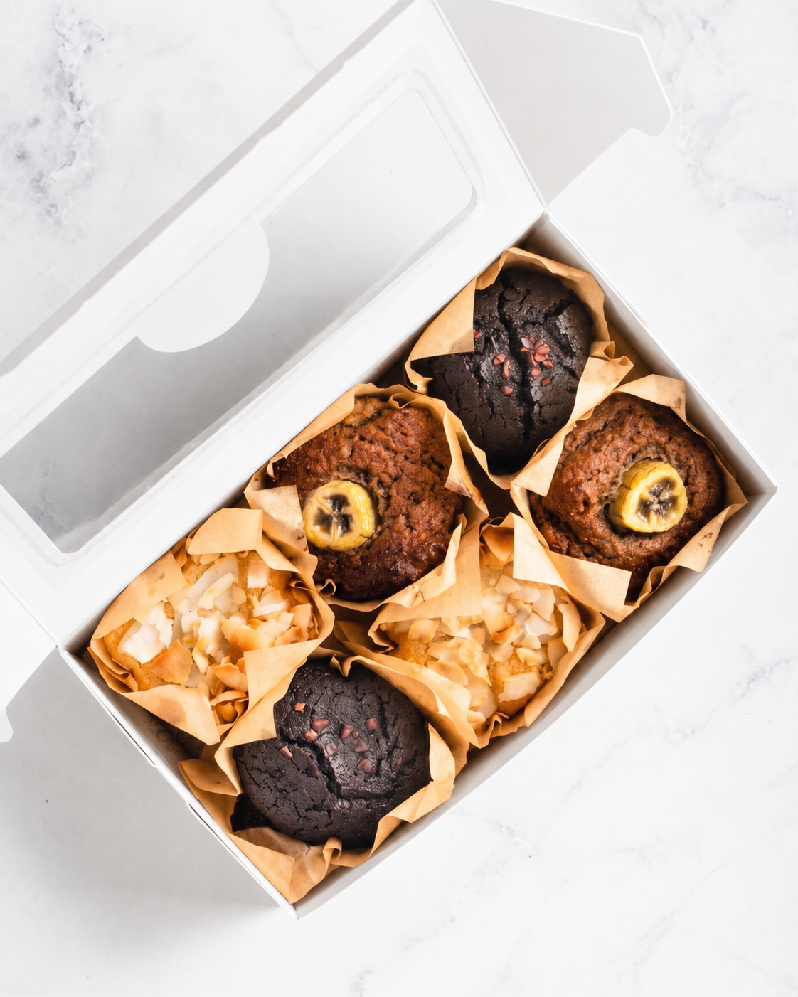 An overhead view of a white bakery box filled with six banana muffins in paper liners, set on a light marble surface.